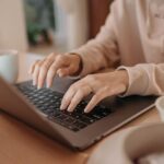 Close-up of a person’s hands typing on a laptop keyboard at a desk, with a coffee mug nearby.