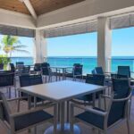 Outdoor beachfront dining area with empty tables and chairs under a covered patio, overlooking turquoise ocean water and palm trees.