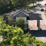Entrance gatehouse with multiple dark metal roofs and two vehicle lanes with barrier arms, surrounded by green trees and landscaping, viewed from above.
